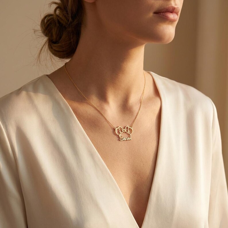 May include: A gold-coloured necklace with a pendant displaying the name "Sarah" in a delicate script. The pendant has a stylized design with two connected shapes above the name. The necklace is worn against a cream-coloured top.