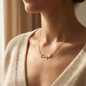 Pu&ograve; includere: Una collana dorata con il nome "Candy" in corsivo. La collana ha una catena delicata e un piccolo ciondolo a forma di cuore. La persona indossa un maglione beige. Un gioiello.