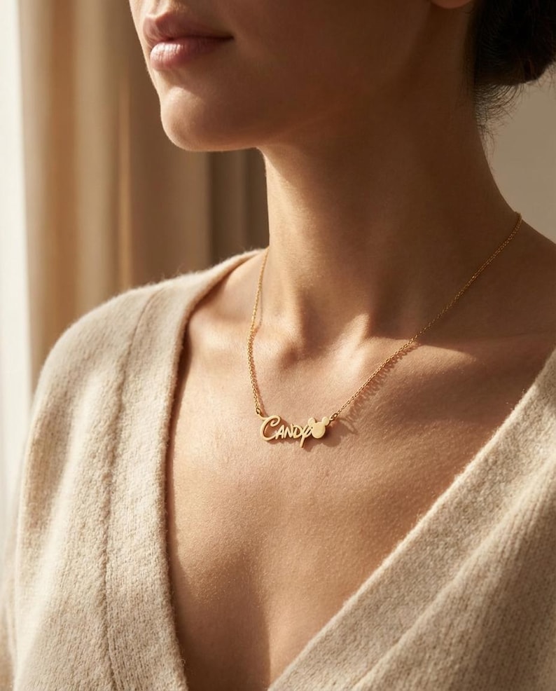 Pu&ograve; includere: Una collana dorata con il nome "Candy" in corsivo. La collana ha una catena delicata e un piccolo ciondolo a forma di cuore. La persona indossa un maglione beige. Un gioiello.