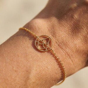 Op de afbeelding: Een goudkleurige armband met een fijne ketting en een cirkelvormige hanger met een geometrisch patroon. De armband wordt om een pols gedragen, wat het elegante ontwerp benadrukt. De hanger heeft een diameter van ongeveer 2 cm.