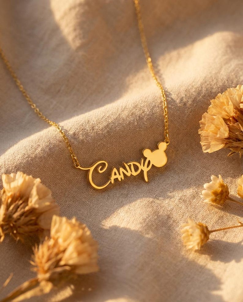 Pu&ograve; includere: Una collana dorata con il nome "Candy" in corsivo, con un design a forma di testa di Mickey Mouse. La collana &egrave; appoggiata su un tessuto beige, con fiori secchi. Il ciondolo e la catena sono dorati.