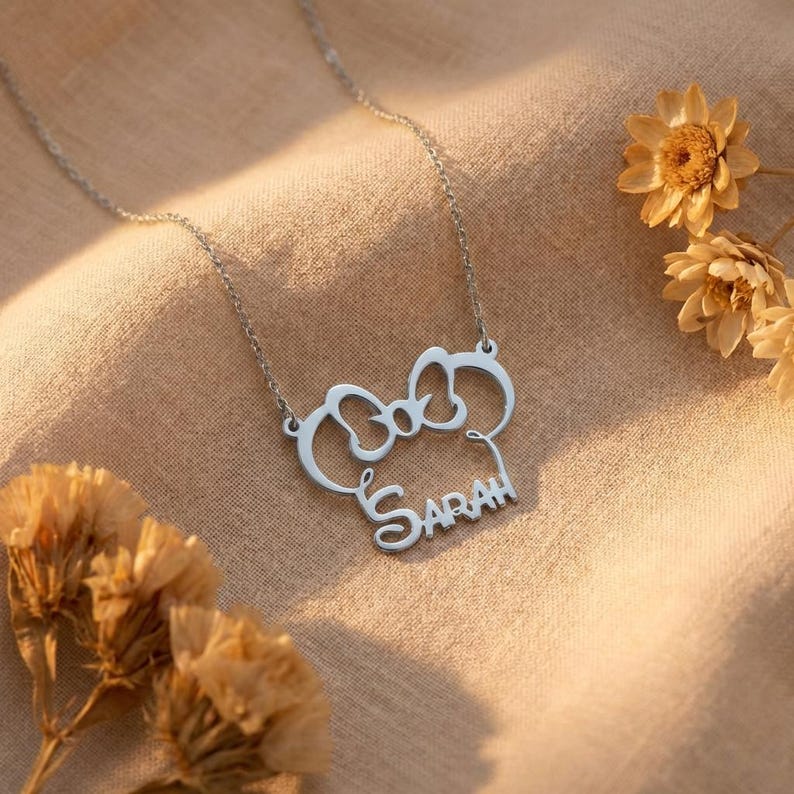 May include: A silver necklace featuring a bow-shaped pendant with the name "SARAH" in a cursive font. The necklace is set against a tan fabric background, with dried flowers in the corners.