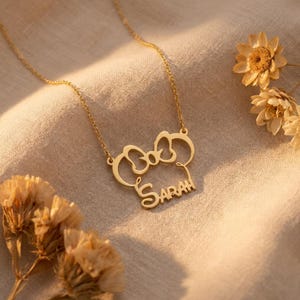 May include: A gold necklace featuring a bow-shaped pendant with the name "Sarah" in a cursive font. The necklace is set against a beige fabric, with dried flowers. The pendant design is a stylized bow, and the chain is fine.