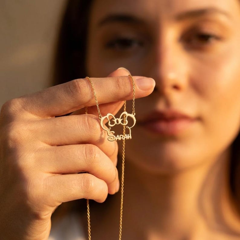 May include: A gold-colored necklace with a bow-shaped pendant and the name "SARAH" in a delicate font. The necklace is held up, showcasing its design and the fine details of the pendant and chain.