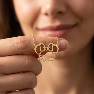 May include: A gold necklace with a bow and a character's head silhouette, with the name "Sarah" in a script font. The pendant hangs from a fine chain, creating a personalised jewellery item.