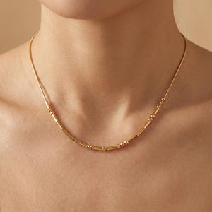 May include: A gold-colored necklace with a delicate chain and a series of small, evenly spaced beads and cylindrical bars. The necklace is worn around the neck, showcasing its simple yet elegant design.