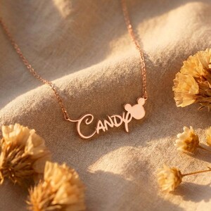 Pu&ograve; includere: Collana in oro rosa con il nome "Candy" in corsivo, ornata da una silhouette della testa di Topolino. La collana &egrave; presentata su un tessuto beige, con fiori secchi in primo piano.