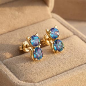 May include: A pair of gold-colored cat-shaped stud earrings. Each earring features a round, blue-purple gemstone for the head and an oval-shaped gemstone for the body. The earrings are set in a gold-colored metal.