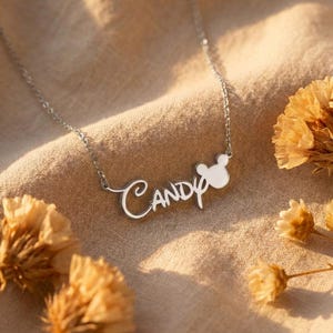 Pu&ograve; includere: Una collana d'argento con il nome "Candy" in corsivo. Il ciondolo presenta un design a testa di topo bianco. La collana &egrave; esposta su uno sfondo di tessuto beige, con fiori secchi.