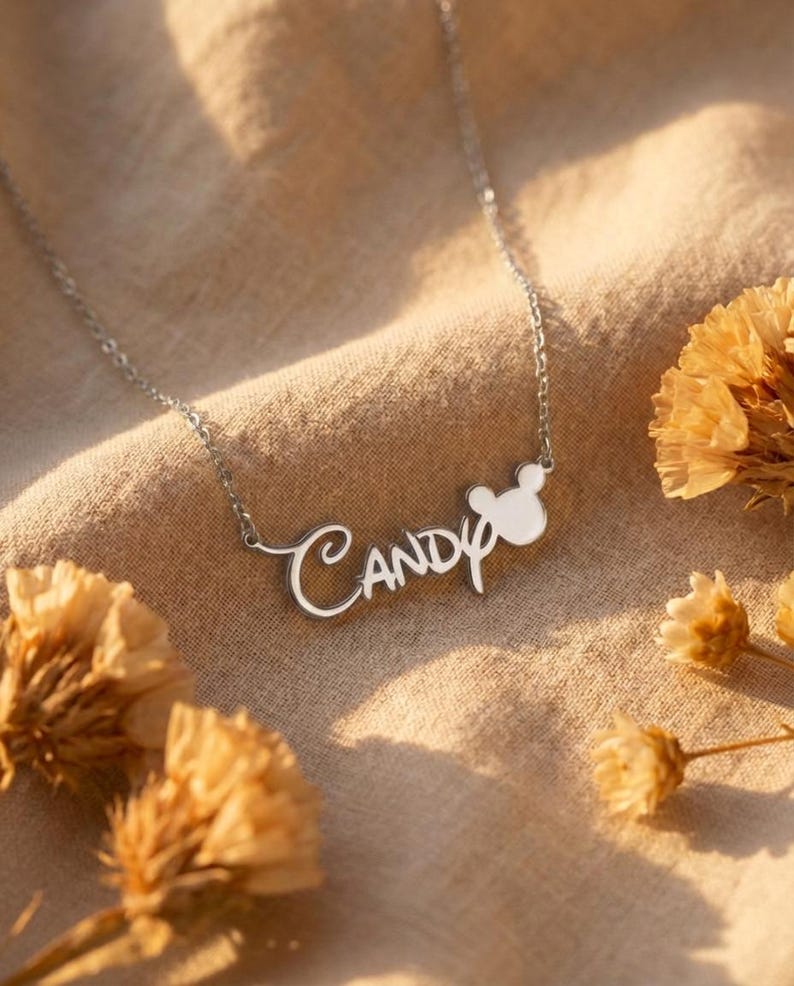 Pu&ograve; includere: Una collana d'argento con il nome "Candy" in corsivo. Il ciondolo presenta un design a testa di topo bianco. La collana &egrave; esposta su uno sfondo di tessuto beige, con fiori secchi.
