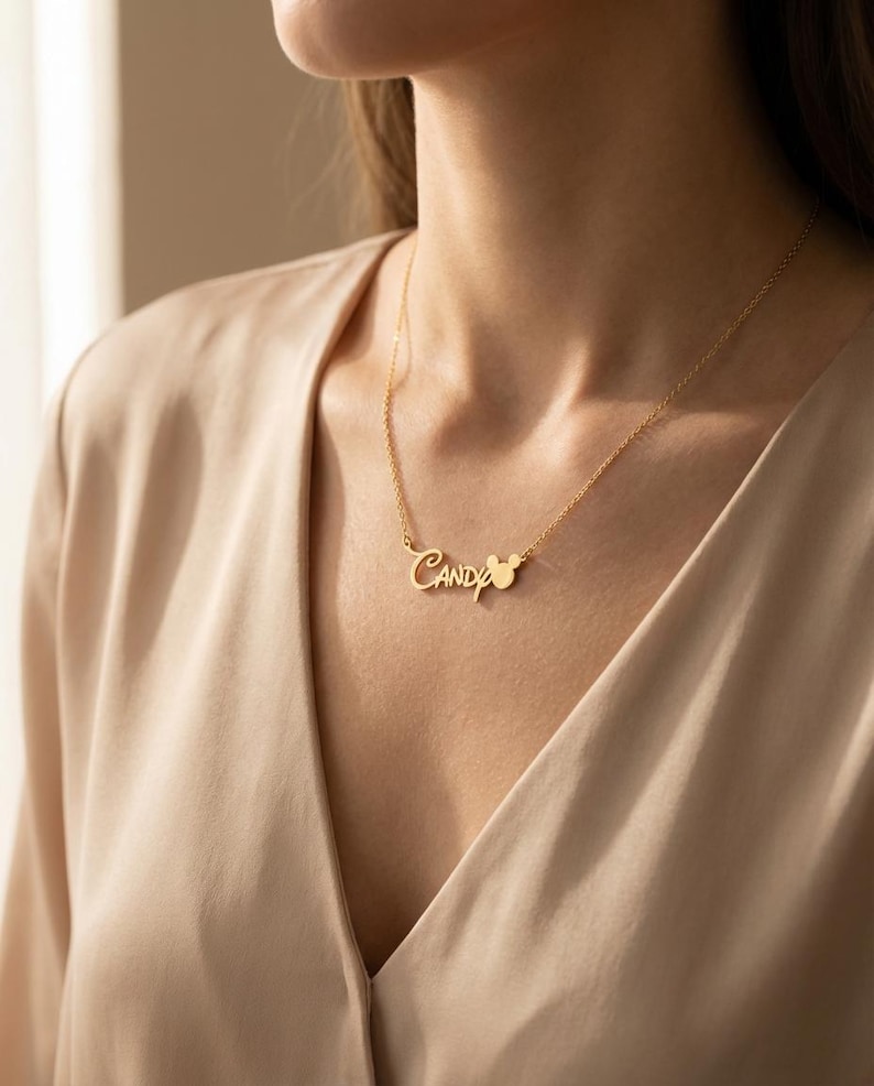 Pu&ograve; includere: Una collana color oro con il nome "Candy" in corsivo. La collana include un piccolo ciondolo a forma di Mickey Mouse. La collana &egrave; indossata da una persona che indossa un top color beige.