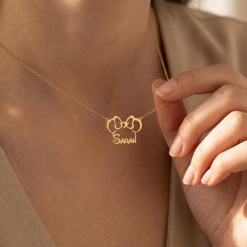 May include: A gold necklace with a pendant shaped like a bow, with the name "Sarah" written in cursive. The necklace is shown against a plain background, emphasizing the intricate design of the jewellery.