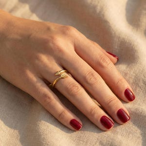 May include: A gold-colored arrow ring. The ring wraps around the finger with the arrow's head and fletching design. The ring is worn on a hand with red nail polish. The background is a neutral fabric.