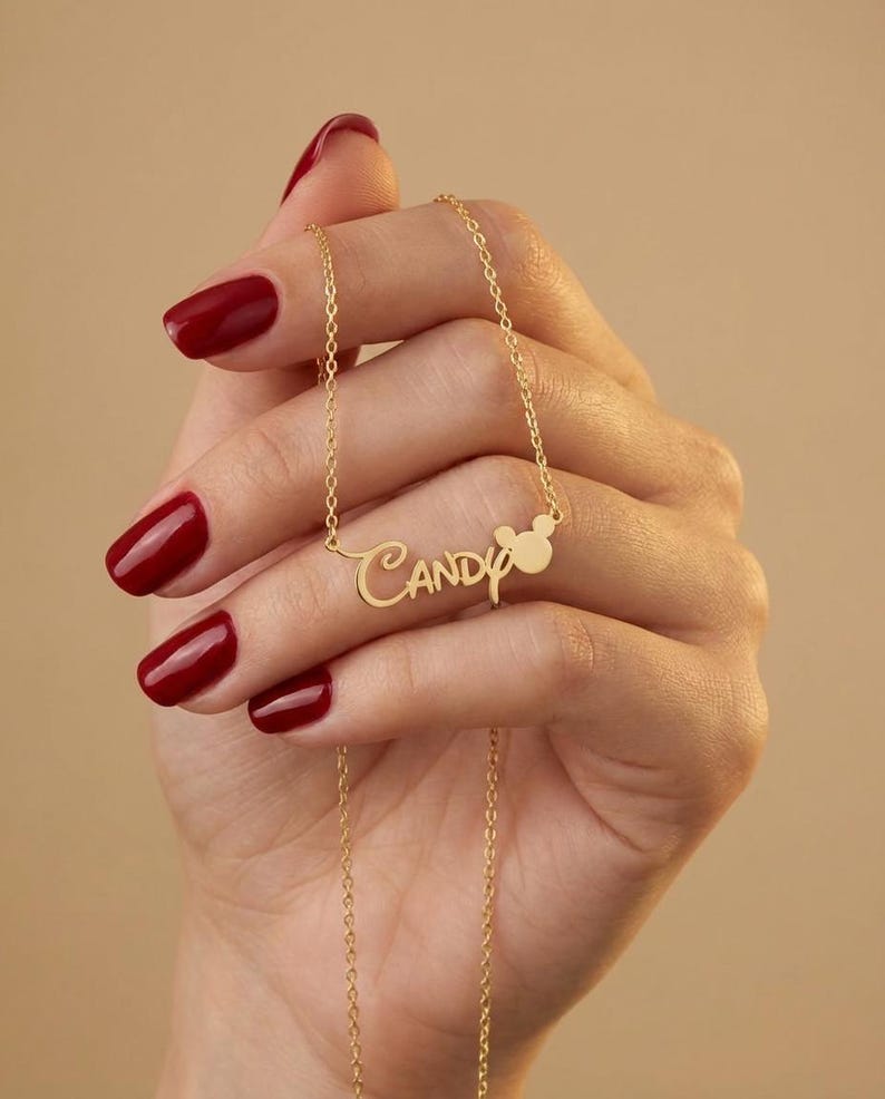 Pu&ograve; includere: Una collana color oro con il nome "Candy" in corsivo. Un piccolo design a forma di testa di Mickey Mouse &egrave; accanto al nome. La collana &egrave; tenuta da una mano con lo smalto rosso.