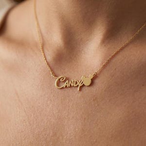 Pu&ograve; includere: Una collana color oro con il nome "Candy" in corsivo. Una piccola testa di Mickey Mouse &egrave; posizionata accanto al nome. La collana &egrave; indossata intorno al collo di una persona.