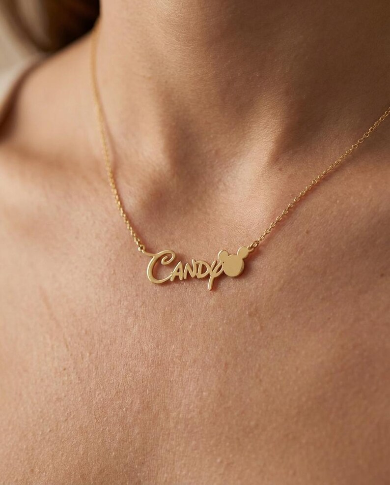 Pu&ograve; includere: Una collana color oro con il nome "Candy" in corsivo. Una piccola testa di Mickey Mouse &egrave; posizionata accanto al nome. La collana &egrave; indossata intorno al collo di una persona.