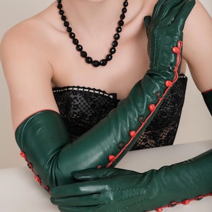 Könnte beinhalten: Smaragdgrüne Opernhandschuhe mit roter Paspelierung und Knöpfen. Die Handschuhe sind aus einem glatten, lederartigen Material gefertigt. Eine schwarze Perlenkette und passende Ohrringe vervollständigen den Look. Die Handschuhe reichen über die Ellbogen hinaus.