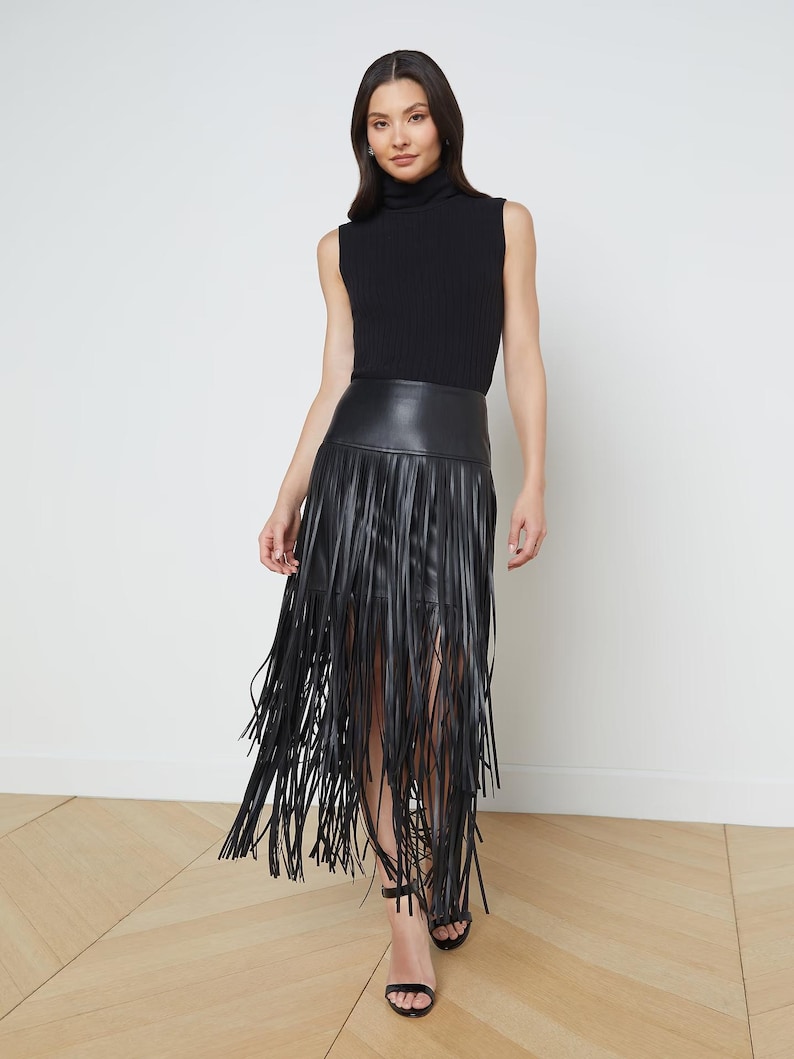May include: A sleeveless black turtleneck top paired with a black leather skirt featuring long fringe. The model is wearing black sandals. The outfit is a stylish ensemble, suitable for a night out or a fashion-forward event.