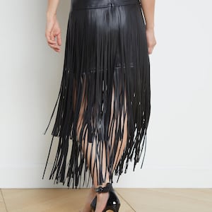 May include: Black faux leather skirt with long fringe detailing. The skirt has a high waist and falls to the mid-calf. The model is wearing black high-heeled sandals. The background is a plain white wall and a wooden floor.