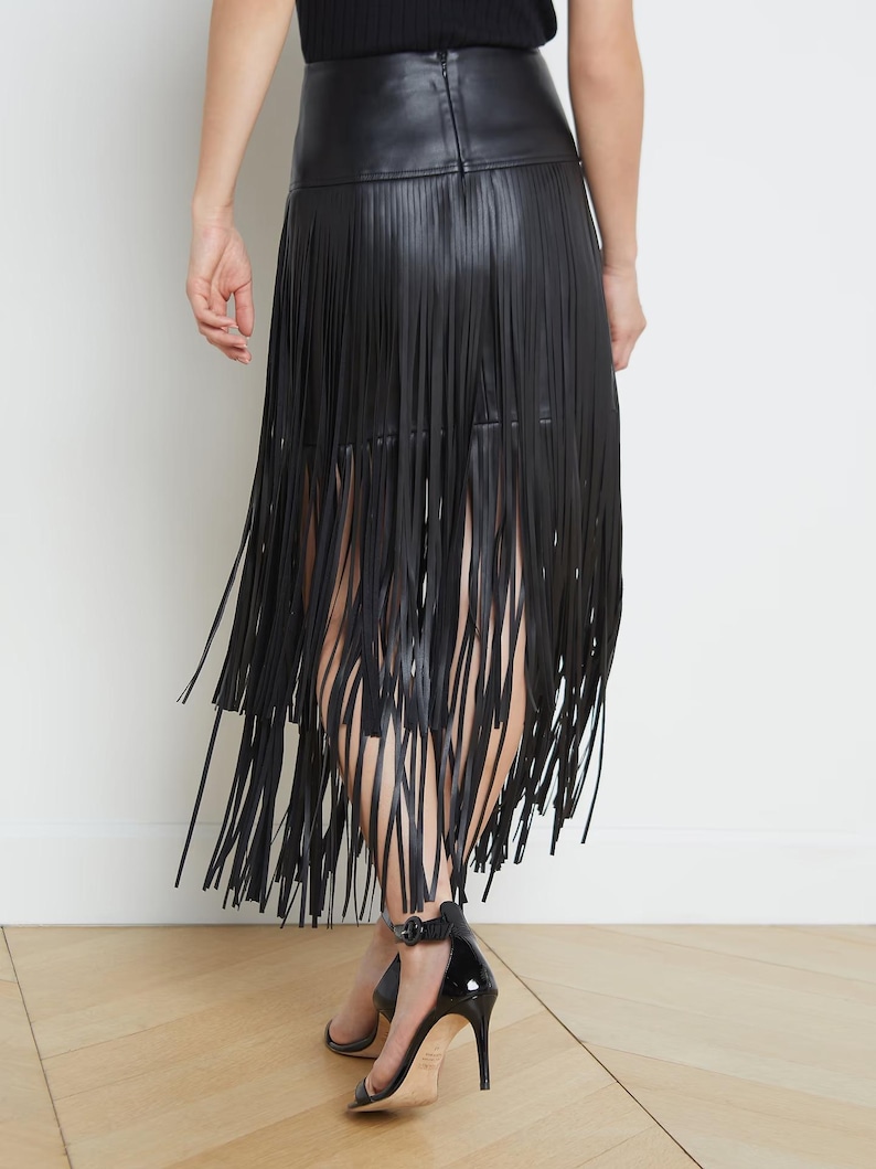 May include: Black faux leather skirt with long fringe detailing. The skirt has a high waist and falls to the mid-calf. The model is wearing black high-heeled sandals. The background is a plain white wall and a wooden floor.