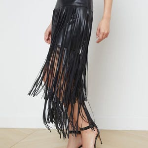 May include: A black leather skirt with long fringe detailing. The skirt has a high waist and falls to the mid-calf. The model is wearing black high-heeled sandals. The skirt is a stylish and edgy fashion statement.