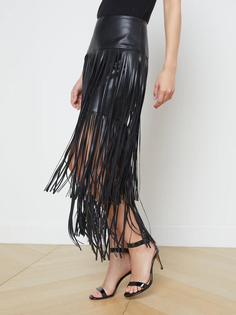 May include: A black leather skirt with long fringe detailing. The skirt has a high waist and falls to the mid-calf. The model is wearing black high-heeled sandals. The skirt is a stylish and edgy fashion statement.