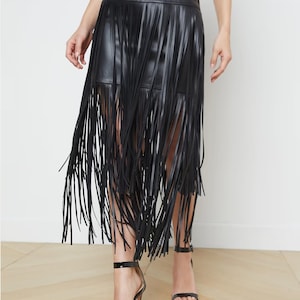 May include: Black faux leather skirt with long fringe detailing. The skirt has a high waist and falls to the mid-calf. The fringe adds movement and texture to the skirt. The model is wearing black high-heeled sandals.