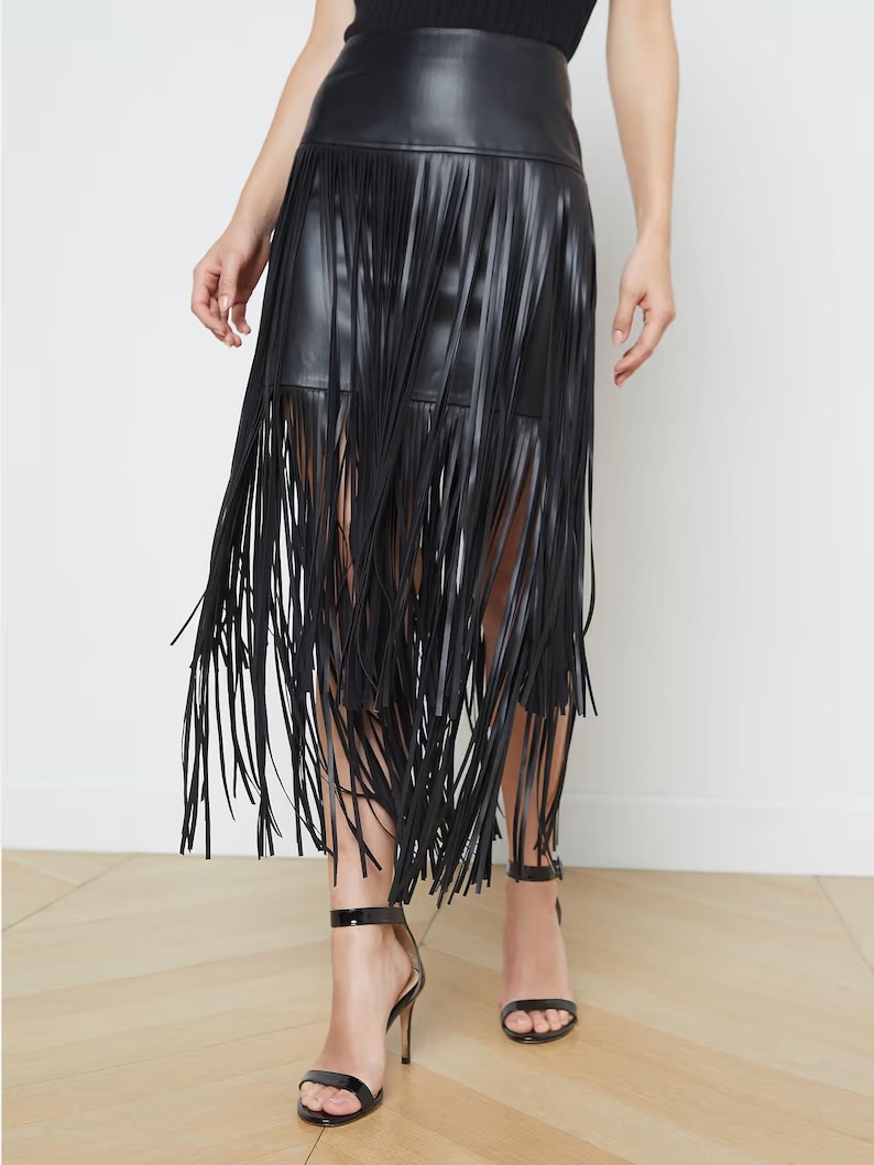 May include: Black faux leather skirt with long fringe detailing. The skirt has a high waist and falls to the mid-calf. The fringe adds movement and texture to the skirt. The model is wearing black high-heeled sandals.