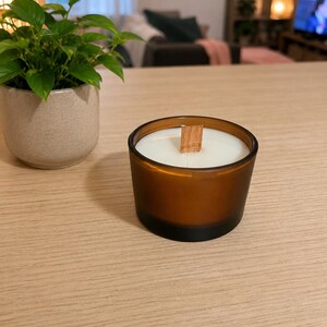 Peut inclure: Une bougie parfumée dans un pot en verre ambré avec une mèche en bois plate centrée dans la cire blanche. La bougie est posée sur une surface en bois clair à côté d'un petit pot en céramique gris moucheté contenant une plante verte.