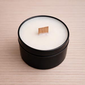 Peut inclure: Une bougie ronde noire remplie de cire blanche et une mèche en bois. La bougie est non parfumée et a un design minimaliste. Le récipient est posé sur une surface en bois de couleur claire.
