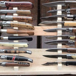 Peut inclure: Une collection de couteaux pliants avec des manches en bois et en os, exposés sur des présentoirs en bois. Les lames en acier varient en forme et en taille. Les couteaux sont disposés en rangées, mettant en valeur leurs différents designs.