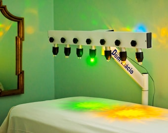 Puede incluir: Un dispositivo de fototerapia horizontal blanco con siete lámparas de casquillo negro montadas en un brazo ajustable. El dispositivo presenta el texto azul Dom Mácio y emite patrones de luz verde y amarilla sobre una superficie blanca.