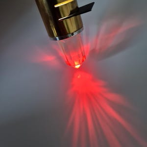 Puede incluir: Una linterna de metal dorado con una punta transparente, facetada y de color rojo brillante. Una pequeña aleta negra está unida al lateral. La linterna emite un haz de luz rojo brillante.