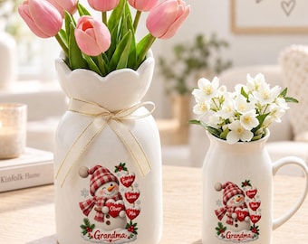 Könnte beinhalten: Eine weiße Keramikvase und ein passender weißer Keramikbecher, beide mit einer Grafik eines Schneemanns mit kariertem Hut und Schal, der ein Herz mit dem Namen Grandma hält. Die Vase enthält rosa Tulpen und der Becher weiße Jasminblüten.