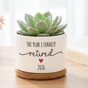 Könnte beinhalten: Ein zylindrischer Übertopf aus weißer Keramik mit einer grünen Sukkulente. Der Topf ist mit dem schwarzen Text "THE YEAR I FINALLY retired", einem kleinen roten Herz und der Jahreszahl "2026" bedruckt. Er steht auf einem runden Holzsockel.