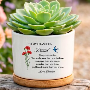 May include: A white ceramic cylindrical planter with a light wood base containing a green succulent. The planter features a red flower illustration, a blue bird, and black text reading: TO MY GRANDSON Daniel Always remember, You are braver than you believe, stronger than you seem, smarter than you think, and loved more than you know. Love, Grandpa.