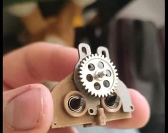 May include: A small, mechanical gear assembly featuring a silver gear with multiple teeth and a series of circular holes. The assembly is mounted on a tan-colored base with two black circular components. The overall design suggests a miniature machine component.