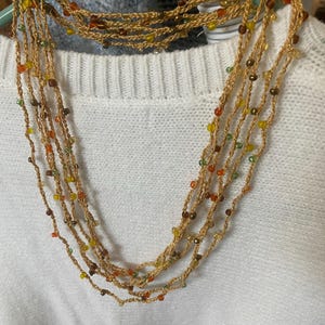 May include: A multi-strand necklace with a gold-colored woven cord and small, colorful beads. The beads are in shades of orange, yellow, green, and brown. The necklace is displayed on a white sweater.