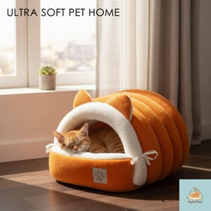 Könnte beinhalten: Ein orangefarbenes Haustierbett mit weißem Innenraum und ohrenförmigen Akzenten. Eine schlafende, rot getigerte Katze liegt in der weichen, geschlossenen Struktur. Der Text "ULTRA SOFT PET HOME" ist oben zu sehen. Ein kleines Pfotenlogo ist vorne sichtbar.