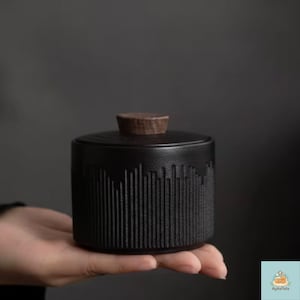 Pode incluir: Um pote cilíndrico de cerâmica preta com um exterior texturizado apresentando linhas verticais de alturas variadas. O pote é coberto por uma tampa circular de madeira castanho-escura que possui um pequeno botão central para manuseio.