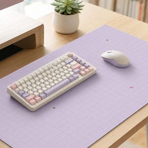 May include: A rectangular light purple desk mat featuring a white grid pattern and small scattered icons. A compact mechanical keyboard with white, pink, and purple keycaps and a matching wireless computer mouse are placed on the mat surface.