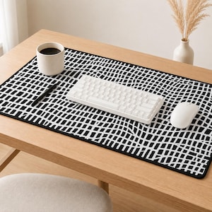 May include: A desk mat featuring a black and white pattern, alongside a white keyboard and mouse. A white mug with coffee and a black pen are also on the desk. The desk is made of light-coloured wood.