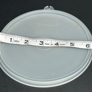以下が含まれることがあります： 透明な丸いTupperwareの蓋。吊り下げ用のループが付いています。白いメジャーで測ると、蓋の直径は約15cmです。Tupperwareのロゴが見えます。