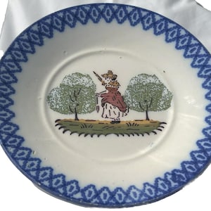 Pode incluir: Prato de cerâmica branco com borda azul estampada. O prato apresenta uma ilustração central de uma pessoa com uma pá entre duas árvores verdes. A pessoa está vestindo um casaco vermelho e um chapéu.