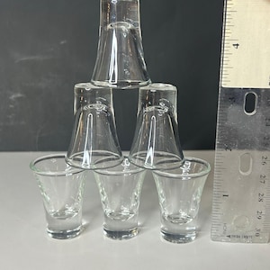 May include: A pyramid of six clear glass shot glasses. The shot glasses are arranged in a triangular formation. A ruler is next to the glasses. The shot glasses are approximately 6 cm tall.