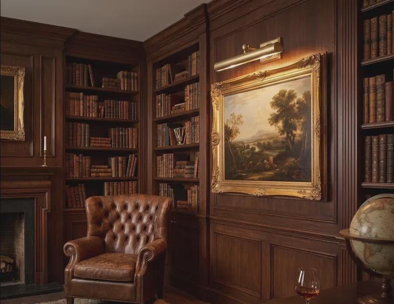 May include: A study room featuring dark wood paneled walls, built-in bookshelves filled with leather-bound books, a tufted brown leather armchair, a landscape painting in a gold frame with a brass picture light, and a globe on a stand.
