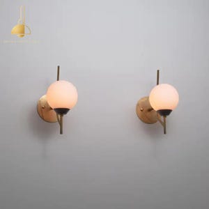 Pode incluir: Duas arandelas de parede com acabamento em latão. Cada arandela apresenta uma lâmpada de globo de vidro fosco. As luzes são montadas em uma haste de latão e uma base circular de latão. O design é minimalista e moderno.