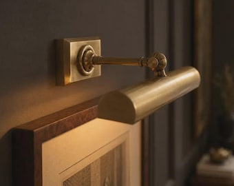 May include: A wall-mounted picture light fixture with a brushed brass finish. The lamp features a rectangular base plate, a horizontal cylindrical shade, and an adjustable arm with a visible hinge mechanism, positioned above a framed artwork.