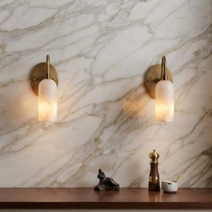 May include: Two wall-mounted sconces featuring cylindrical, translucent white glass shades and brushed brass hardware. The fixtures emit a warm, soft glow, positioned symmetrically against a white marble surface with grey veining.