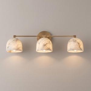 May include: A three-light vanity wall fixture featuring a horizontal brass bar and a circular brass wall mount. Three dome-shaped shades with a white and grey marbled pattern are attached to the bar, each housing a single light bulb.